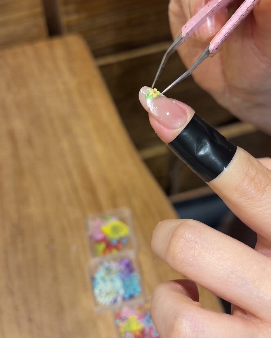 Precision tweezers placing a tiny yellow dried flower onto the wet gel surface of a fingernail.