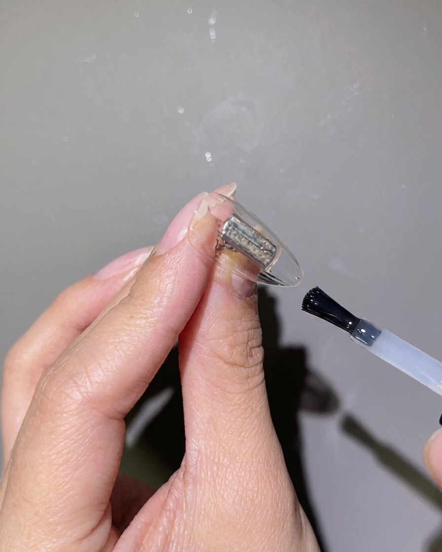 A close-up of a nail brush applying clear reinforcing gel over a clear nail tip.