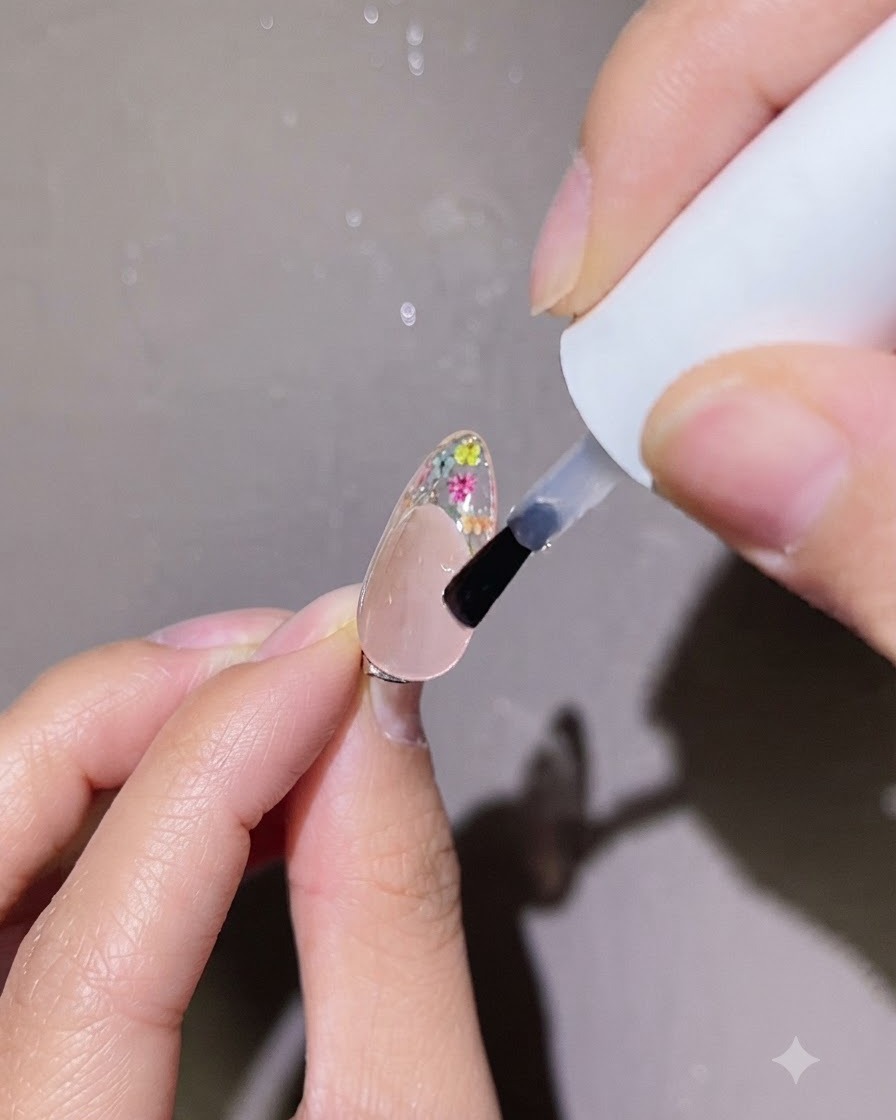 A small brush applying a precise dot of clear top coat onto the floral nail art on a fingernail.