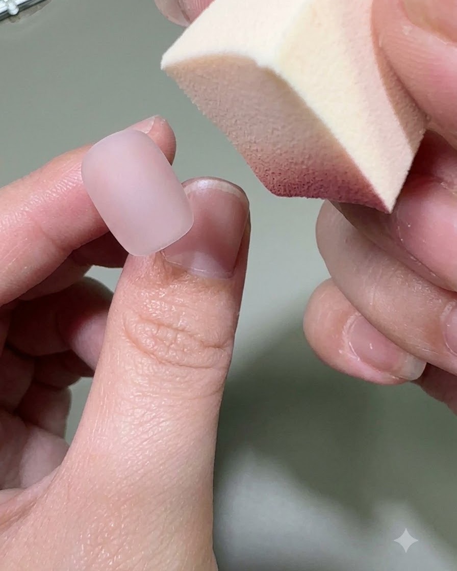 A hand holding a makeup sponge applying pink gel color onto the tip of a matte nude nail.