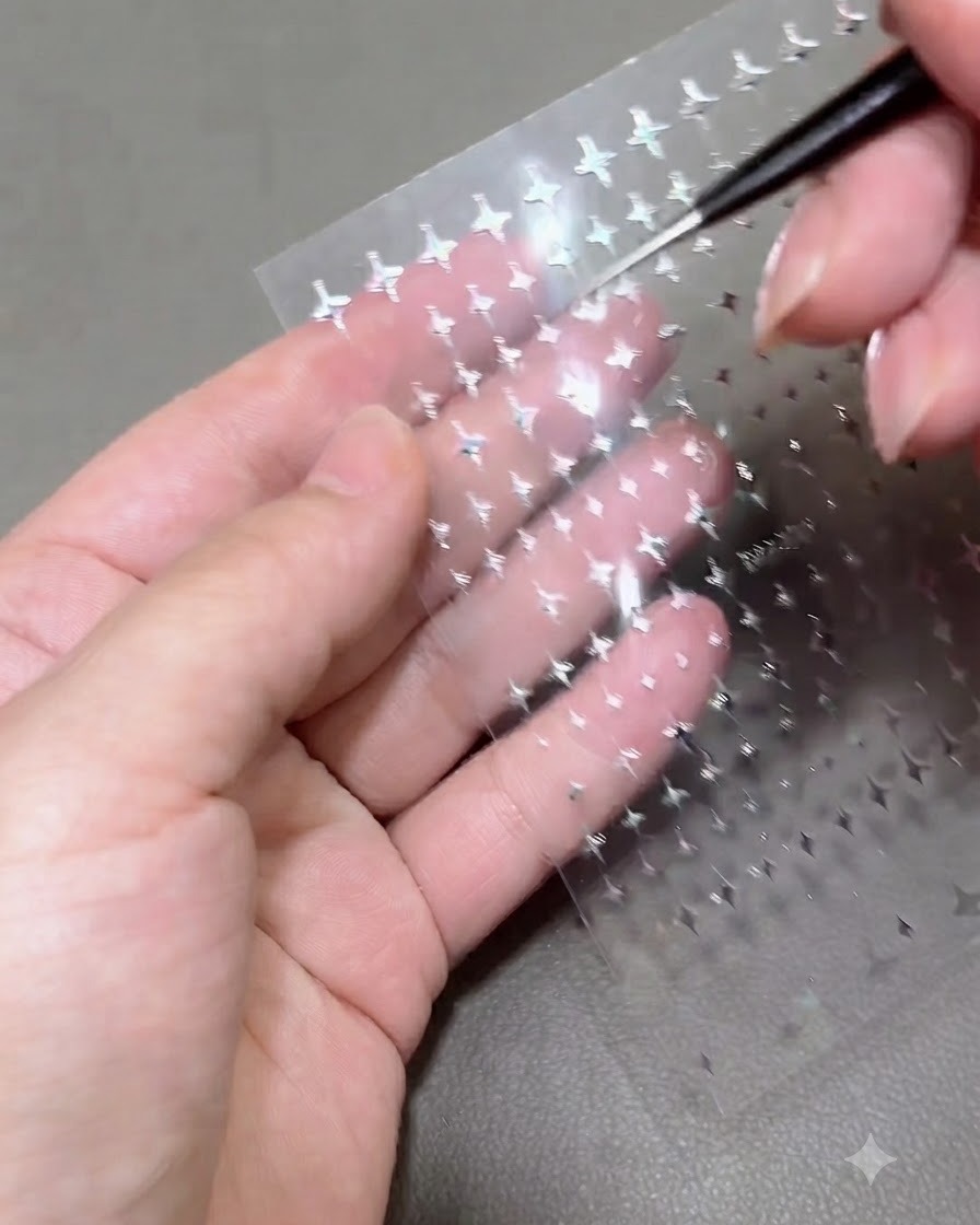 A close-up of a detailing tool lifting a tiny silver star sticker from a transparent sticker sheet held over a persons palm.