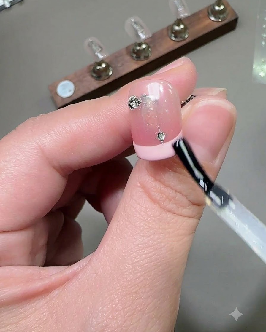 A brush applying a glossy top coat layer over a decorated nail with rhinestones and star charms.