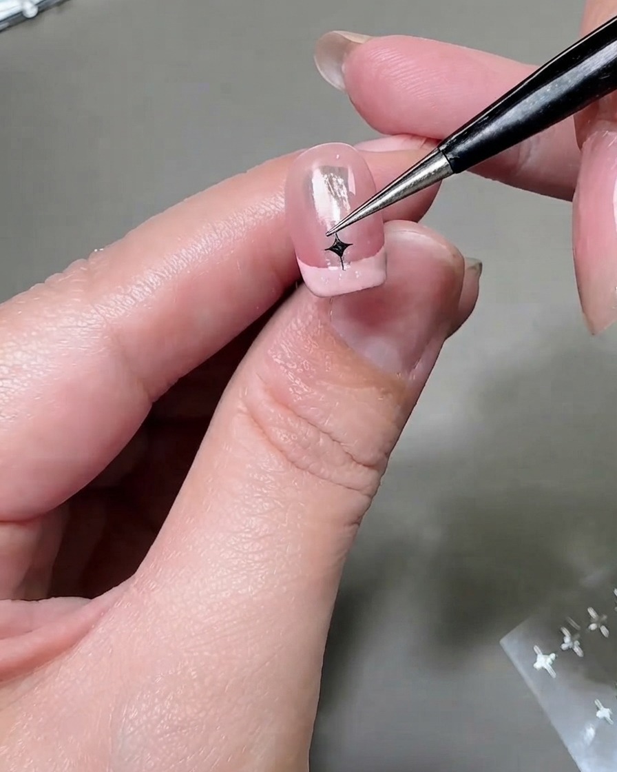 A fine tool positioning a silver star sticker on a second French manicured nail tip.