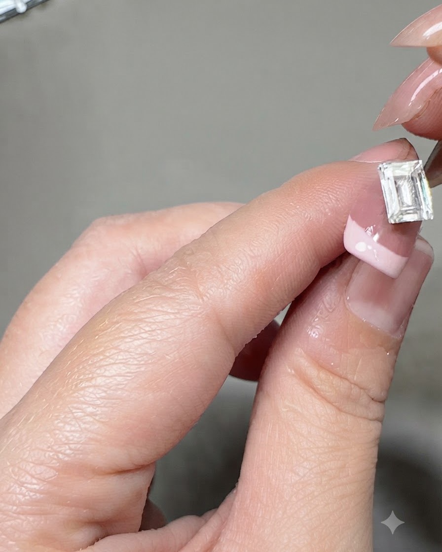 A pair of tweezers placing a large rectangular crystal gem onto the center of a French manicured nail.