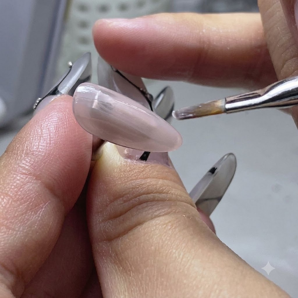 A flat brush applying a thin coat of nude color base gel onto a practice nail finger.