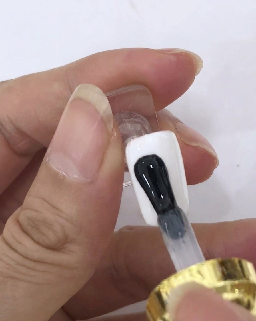 A brush applying a clear coat of blooming gel onto a white-painted nail surface.