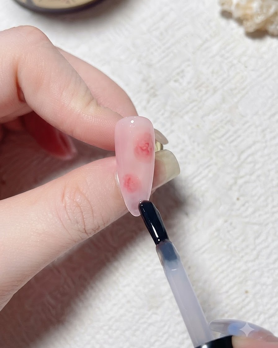 A brush applying a clear layer of blooming gel onto a pink-toned artificial nail.