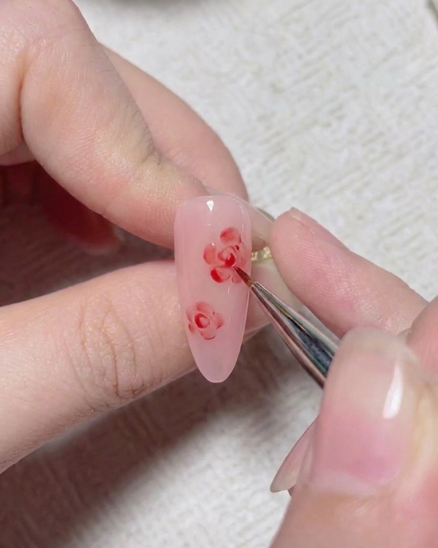 A fine nail art brush adding red detail lines to form the center of a painted rose on a pink nail.