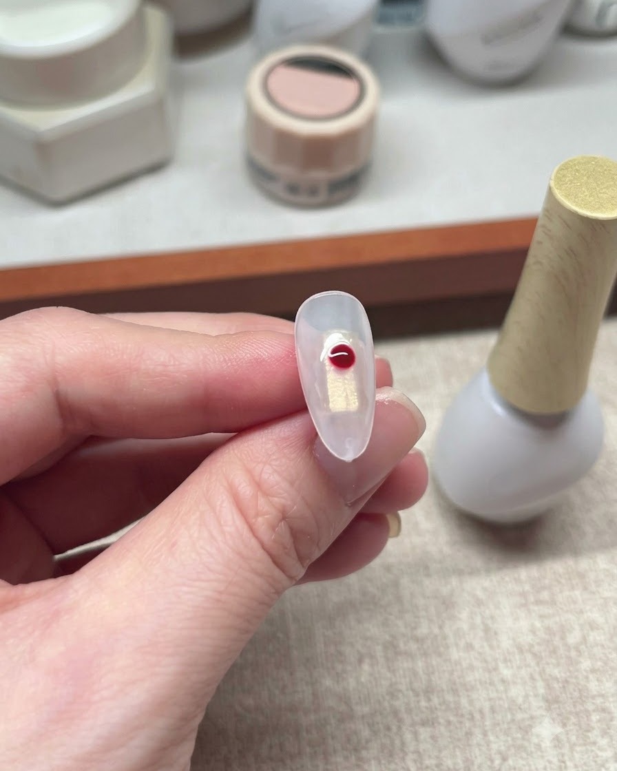 A single red dot of gel slightly blooming on a clear nail surface.