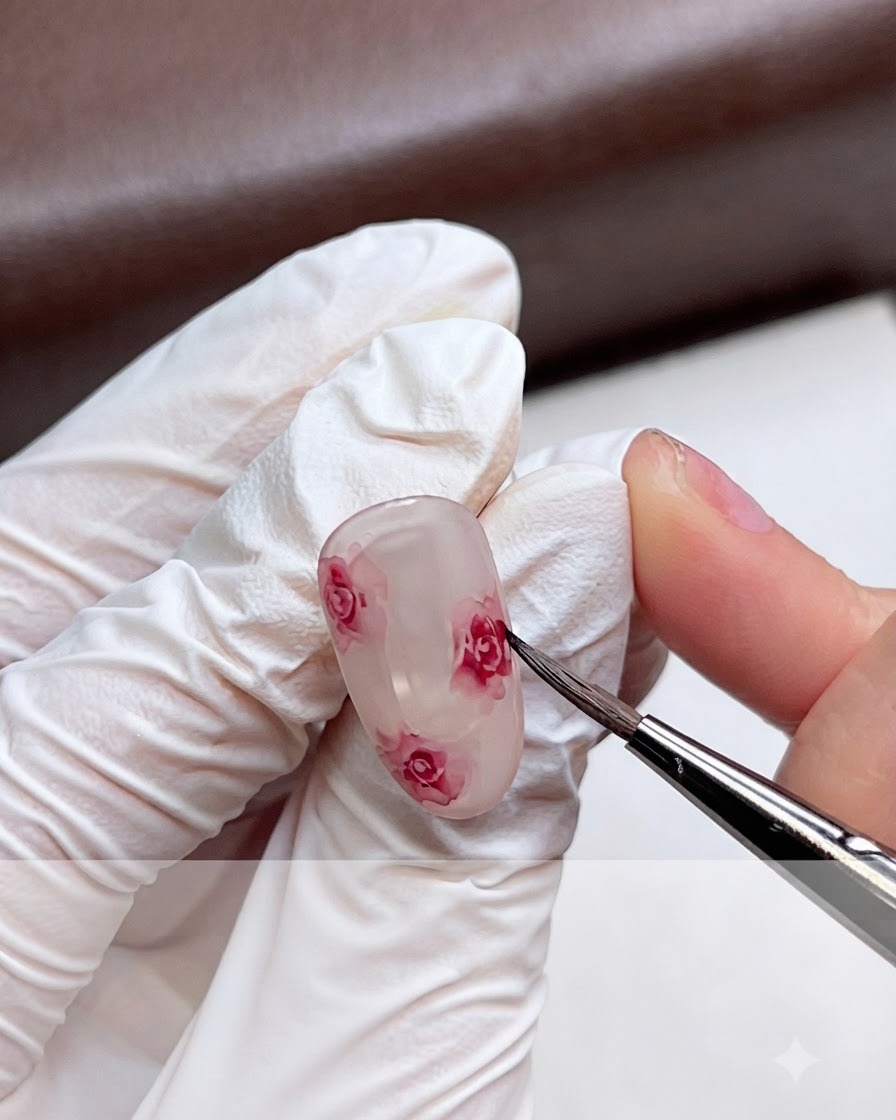 A gloved hand gently blending the edges of a pink watercolor rose on a gel nail using a fine brush.