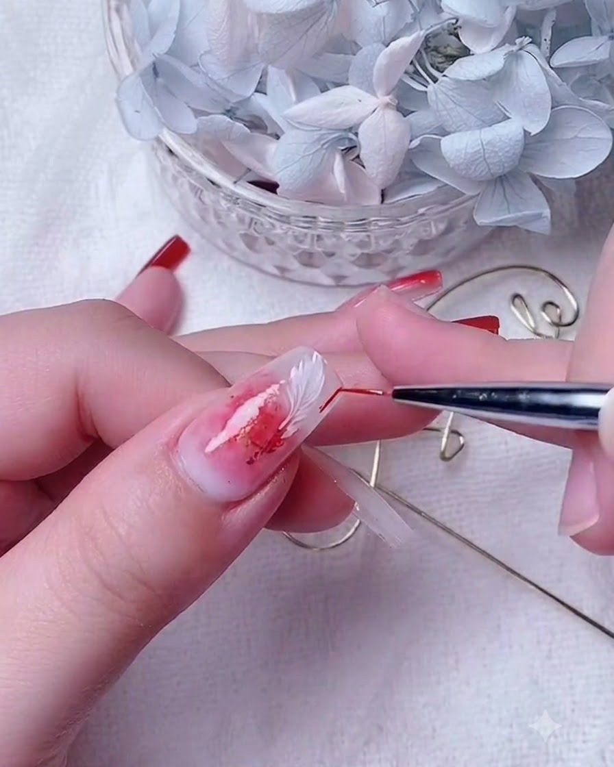 A close-up of a nail artist using a thin detail brush to blend red gel on a milky white base nail.