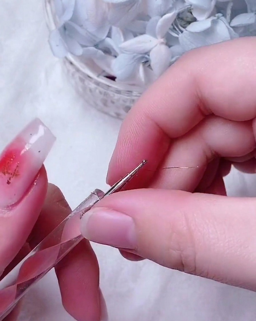 Fine tweezers placing a small, intricate gold wire rose onto the center of a decorated fingernail.