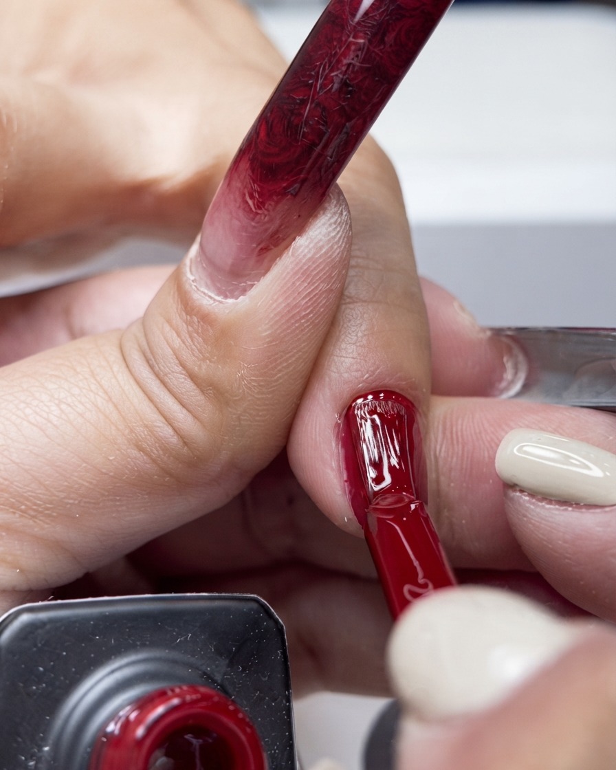 A brush applying a vibrant red gel polish down the center of a fingernail.