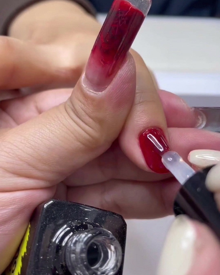A close-up of a nail brush applying clear top coat over a glossy red gel manicure.