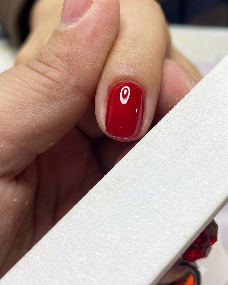 A white nail file gently shaping and smoothing the edge of a completed red gel nail.
