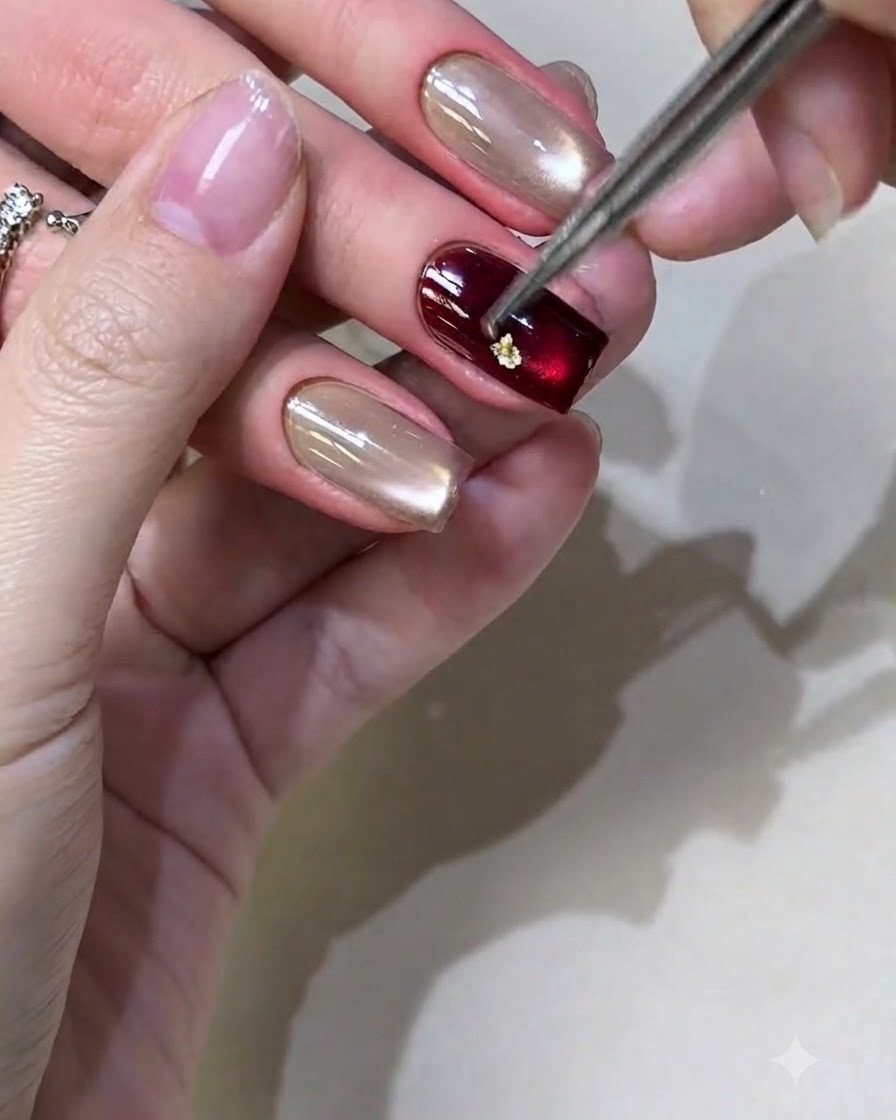 A pair of tweezers placing small pieces of gold foil onto a burgundy gel polished nail.