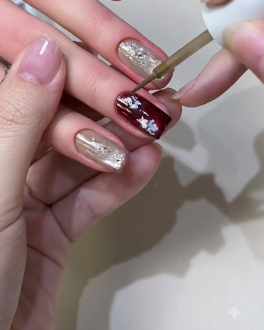 A fine nail art brush applying metallic liquid to form small casual flower patterns on a burgundy nail.
