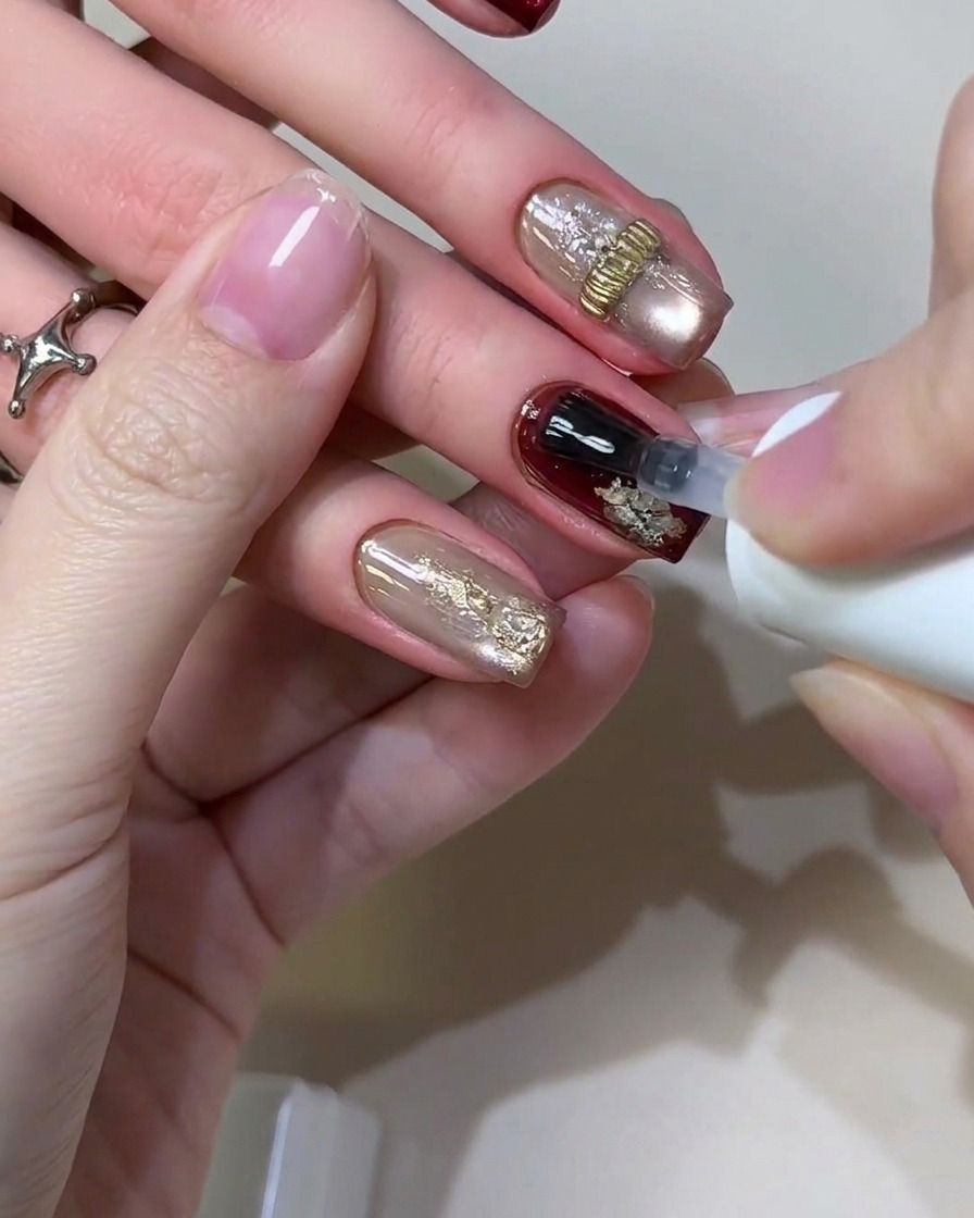 A gel brush applying a clear glossy top coat sealant over a completed retro burgundy and gold foil nail design.