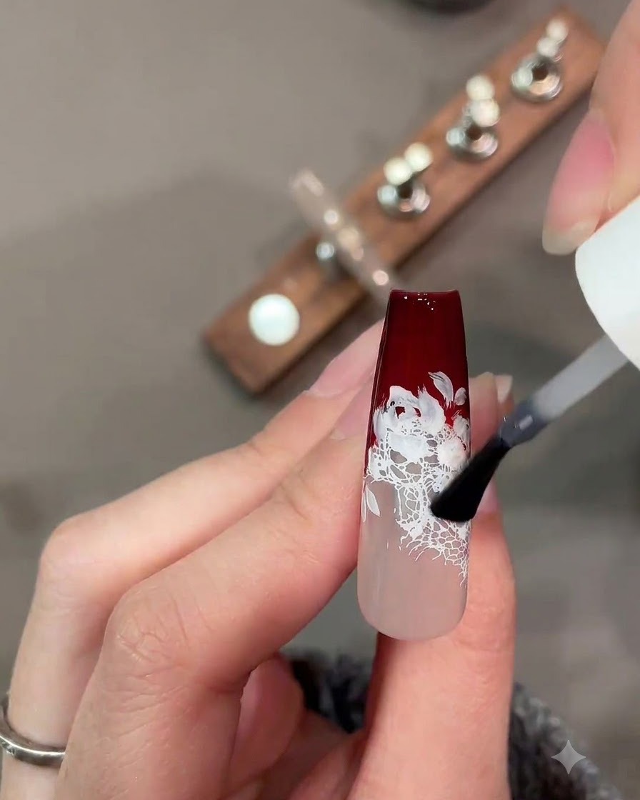 A brush applying a matte top coat over a dark red and white lace nail design.