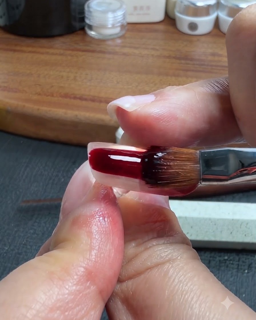 A nail brush applying a dark red-brown translucent gel onto the lower portion of a clear artificial nail.