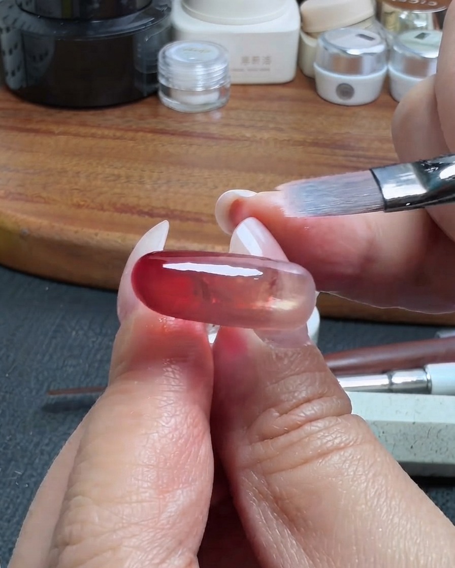 A clean makeup brush gently blending the edge of a red translucent gel layer on a nail.