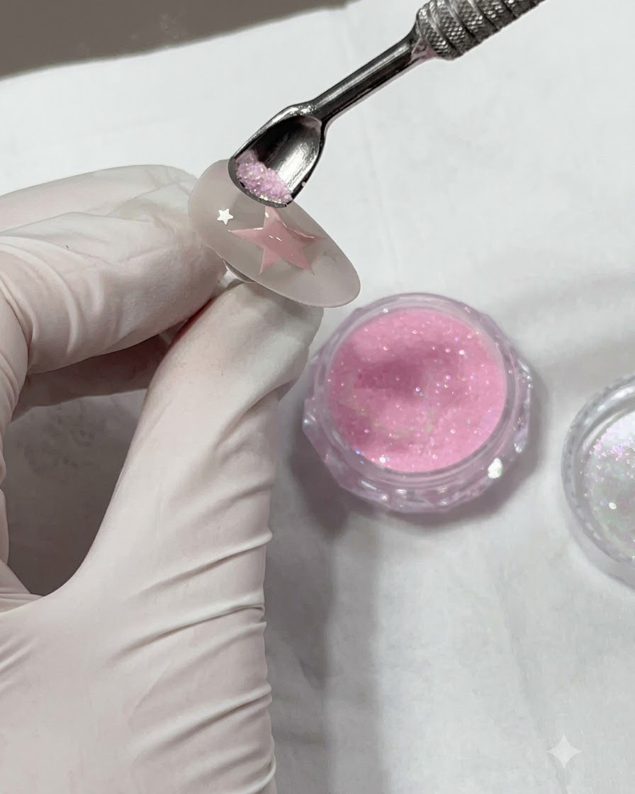 A metal cuticle pusher holding pink glitter powder above a painted star on a nail.