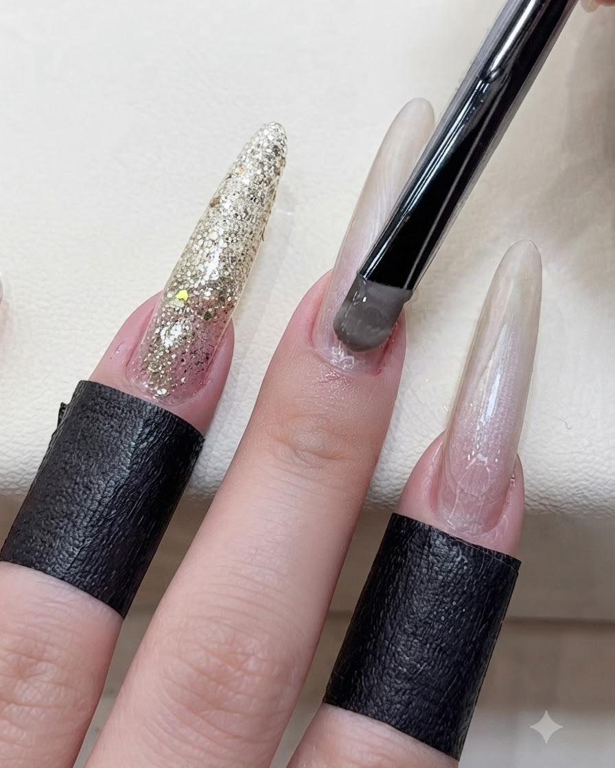 A close-up of a hand using a black-handled brush to apply greyish-brown cat eye gel onto the nail bed of an extended finger.