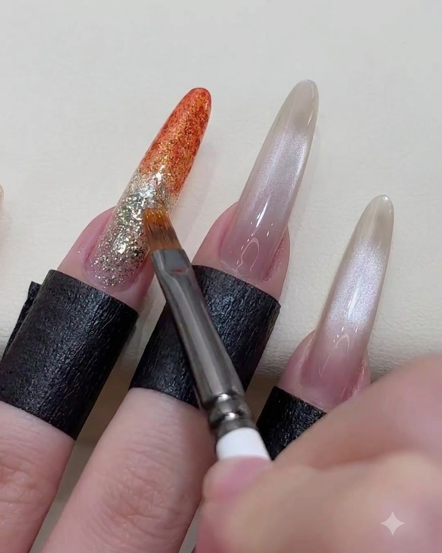 A close-up view of a hand using a fine brush to feather and blend the transition line of a vibrant orange gel painted over a glitter base layer.