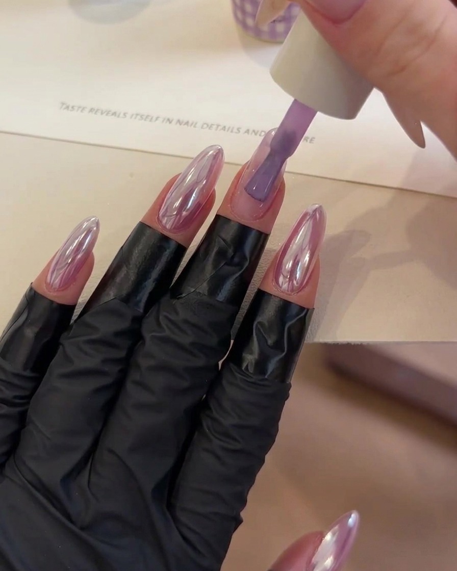 A brush placing a dollop of clear building gel onto the center of a purple-toned fingernail.