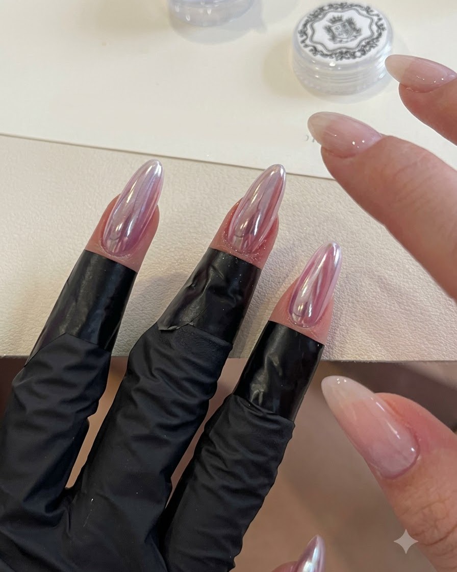 A sponge applicator buffing iridescent sky mirror powder onto almond-shaped chrome nails.