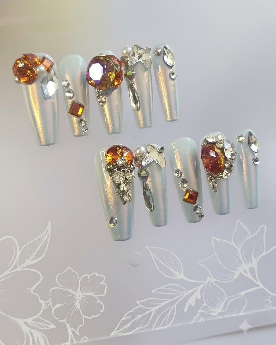 A macro shot focusing on feature press-on nails with amber gradient backgrounds and clusters of large sparkling jewel stones.