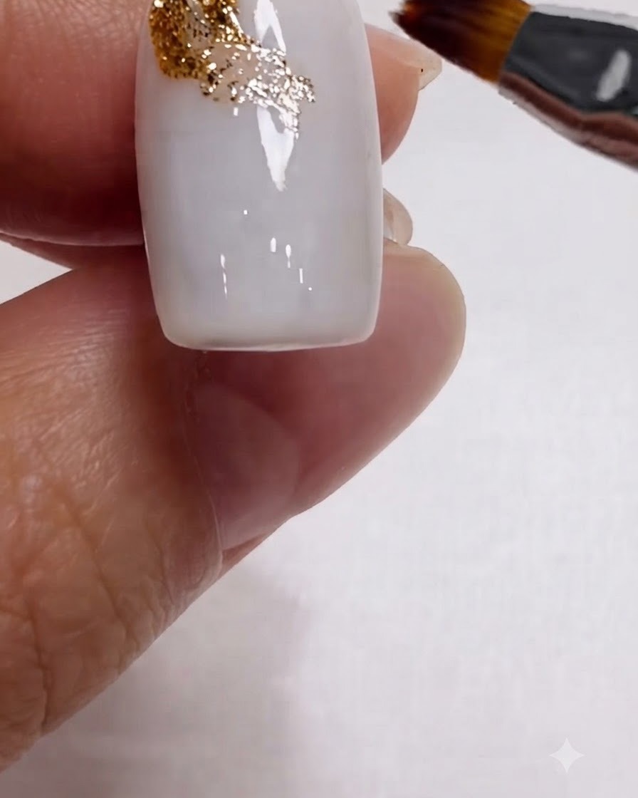 A brush blending golden glitter diagonally across the upper corner of a milky white nail.
