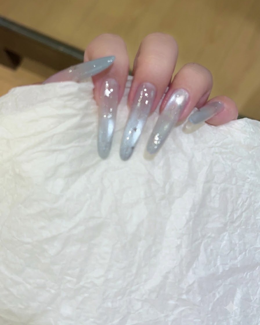 A hand holding a white textured tissue wiping down long, translucent gel-extended nails.