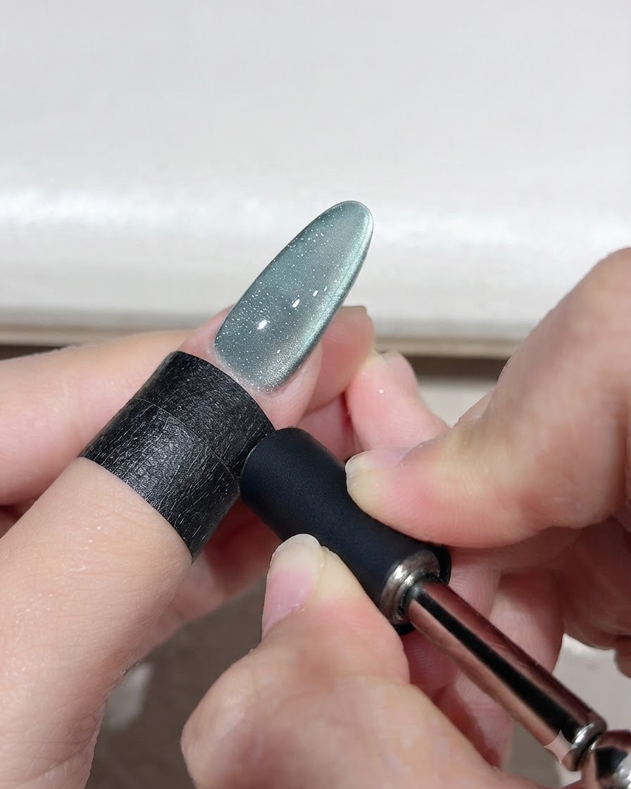 A magnetic tool held near a green cat-eye gel nail to shape a curved light arc.