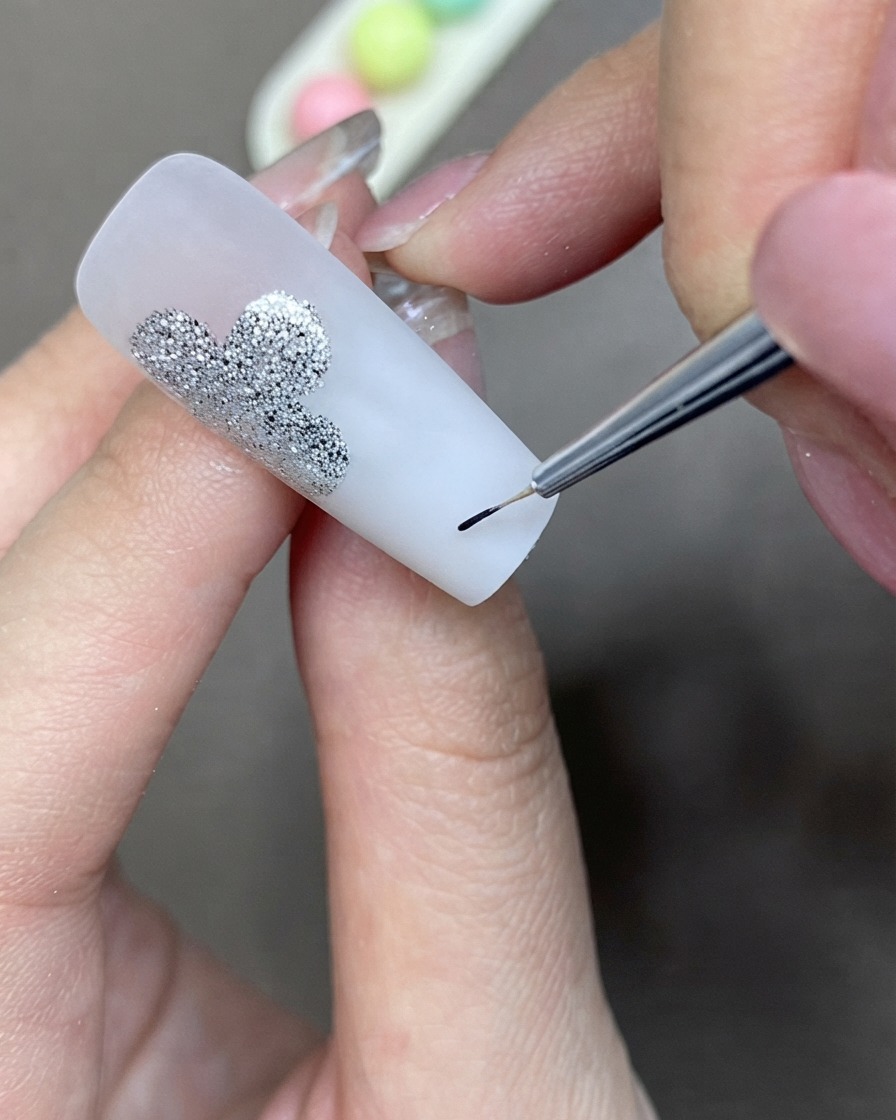 A fine detail brush applying black liner gel to create a clover shape next to a silver glitter design.