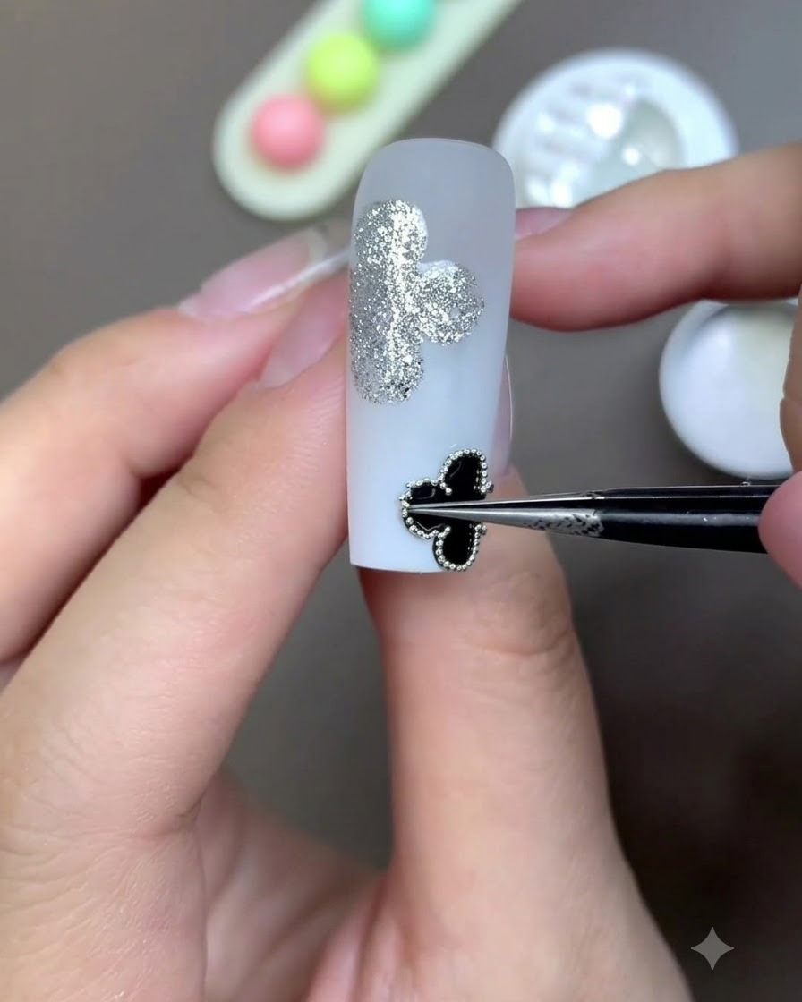 A pair of precision tweezers placing a framed black clover motif onto the surface of a milky white nail.