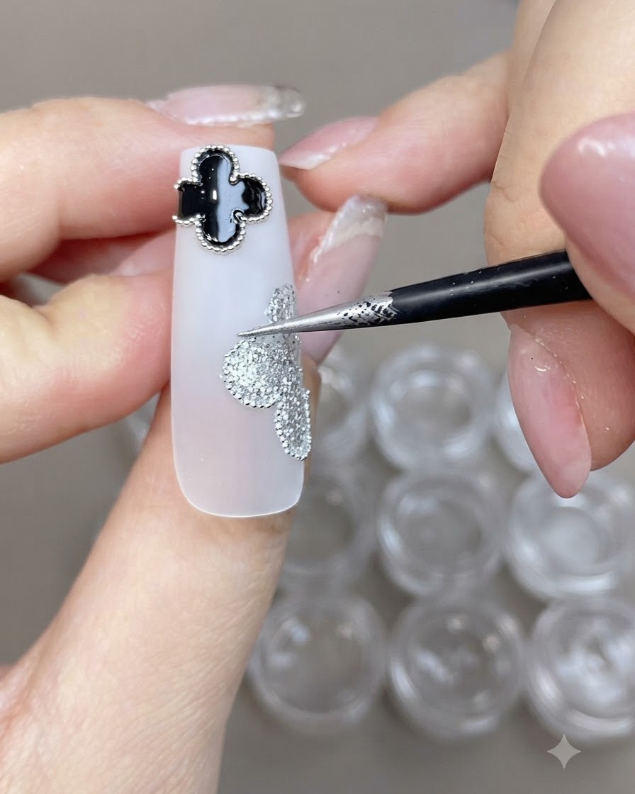 A fine brush placing small steel beads along the edge of a silver glitter clover design on a white nail.