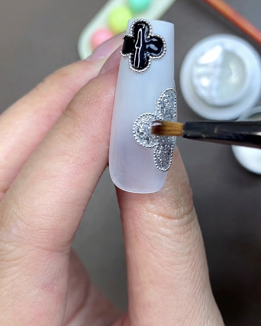 A brush applying clear rhinestone adhesive gel to the center of a silver glitter clover motif on a nail.