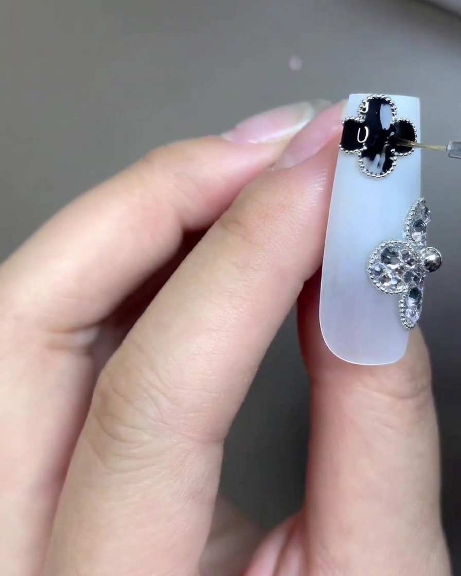 A close-up of a long milky white nail adorned with two four-leaf clover charms—one black, one made of rhinestones. A very fine nail art brush is applying clear top coat precisely onto the black clover detail.