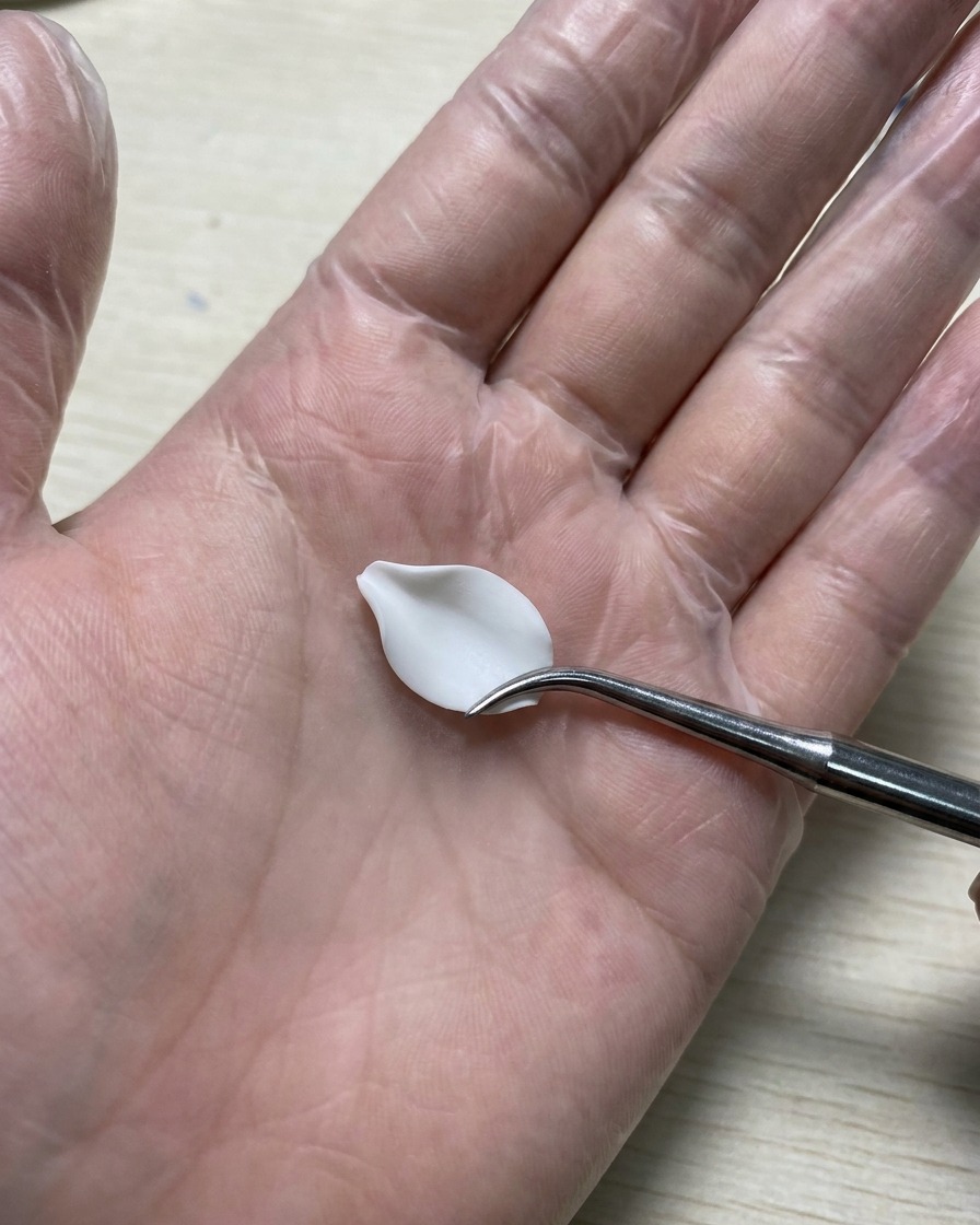 A gloved hand holding a silicone carving tool smoothing out a white 3D gel petal resting on a silicone mat.