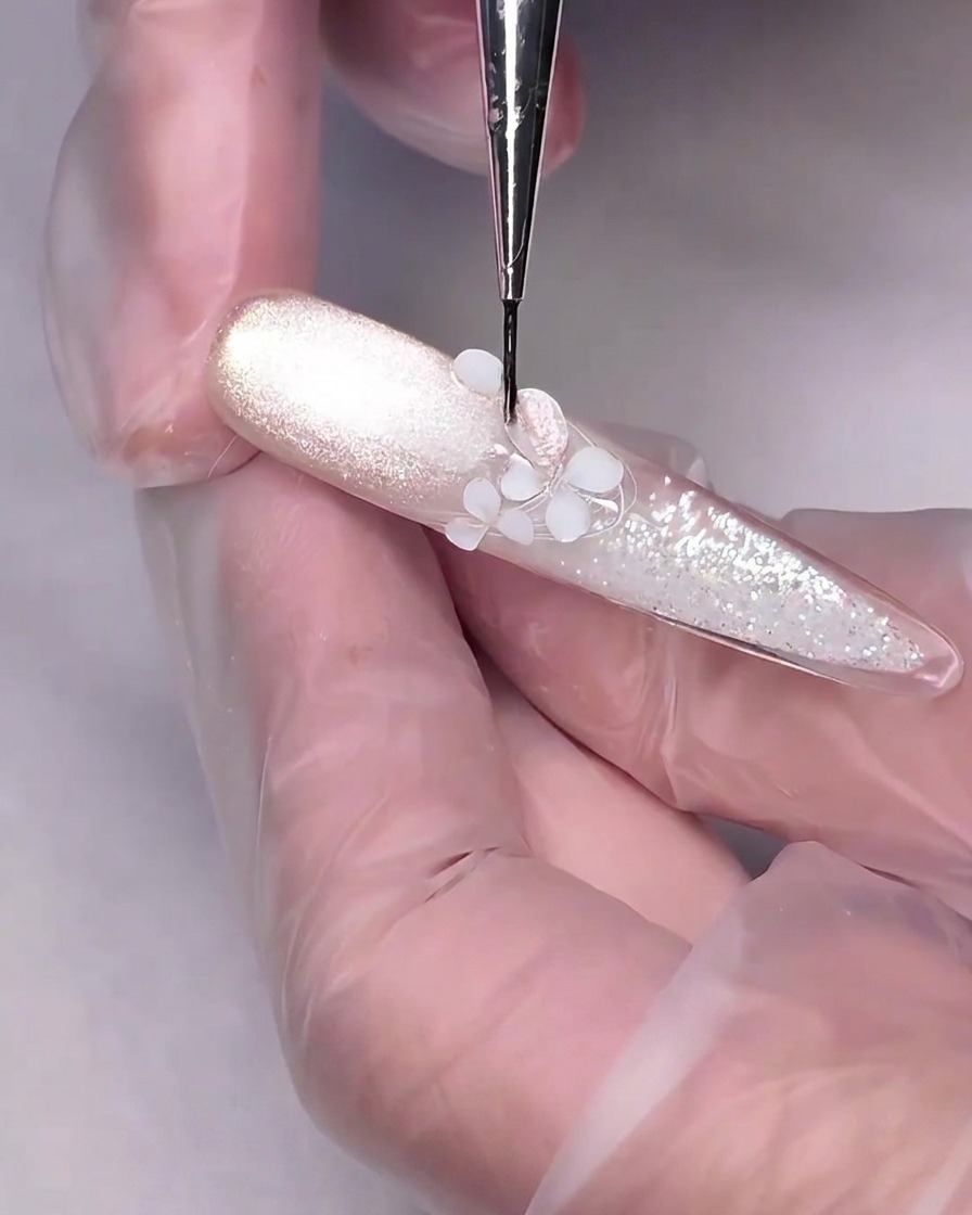 A fine detailing brush applying nude-pink cat-eye gel onto a white 3D sculpted flower on a nail.