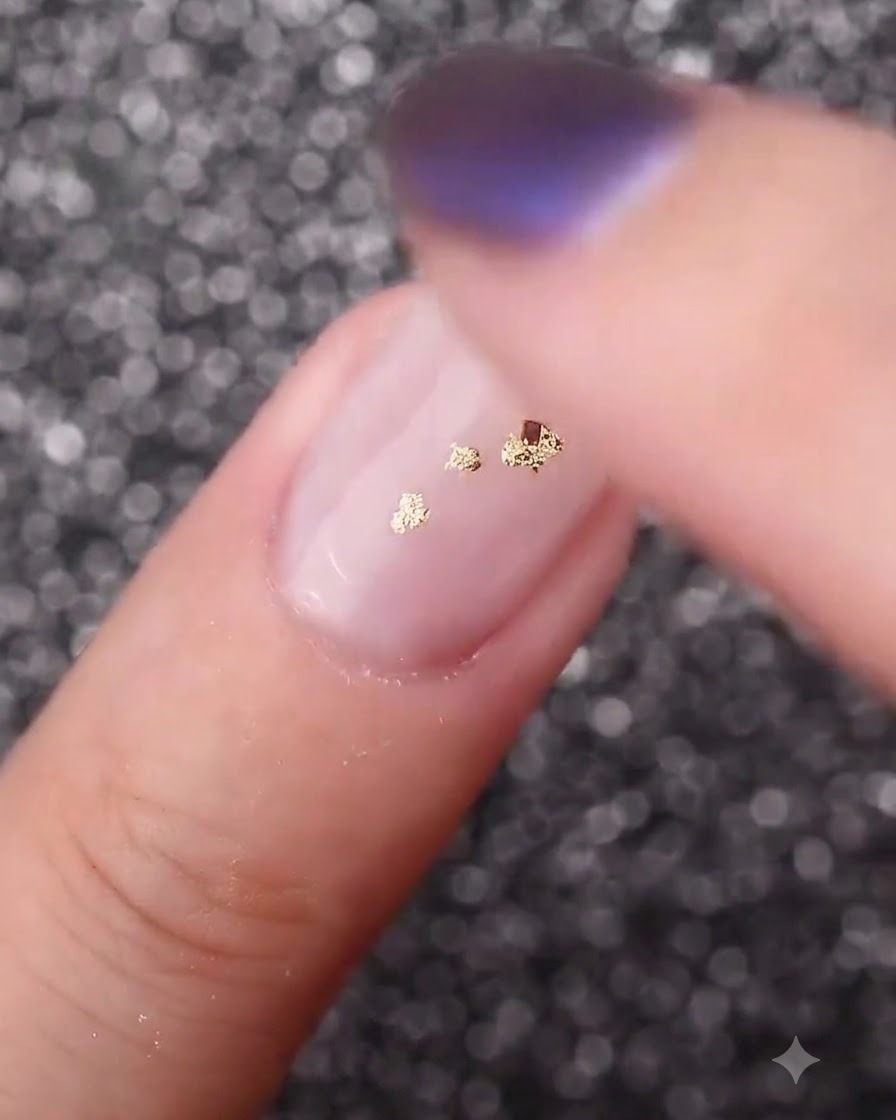 A finger gently pressing down on gold foil flakes to secure them flat against the curved nail surface.