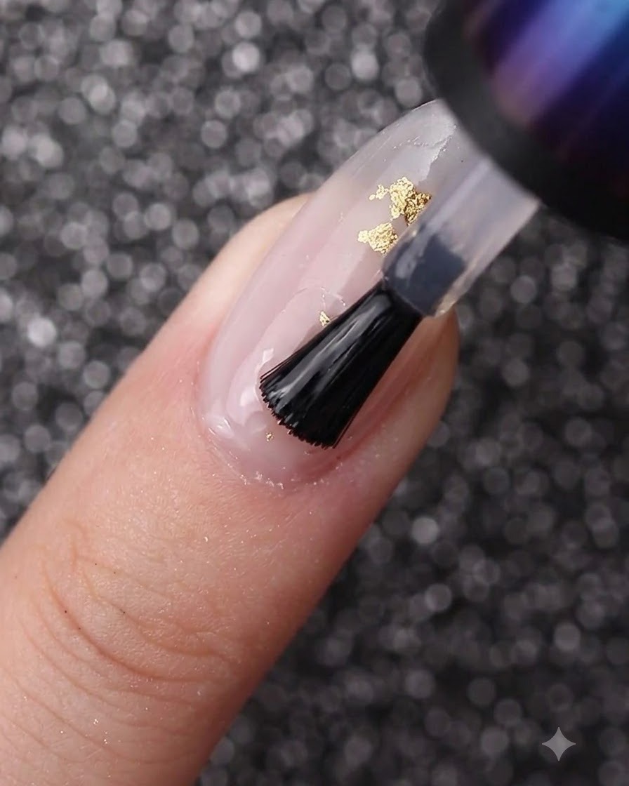 A nail brush spreading clear gel over a nail decorated with milky white polish and gold foil.
