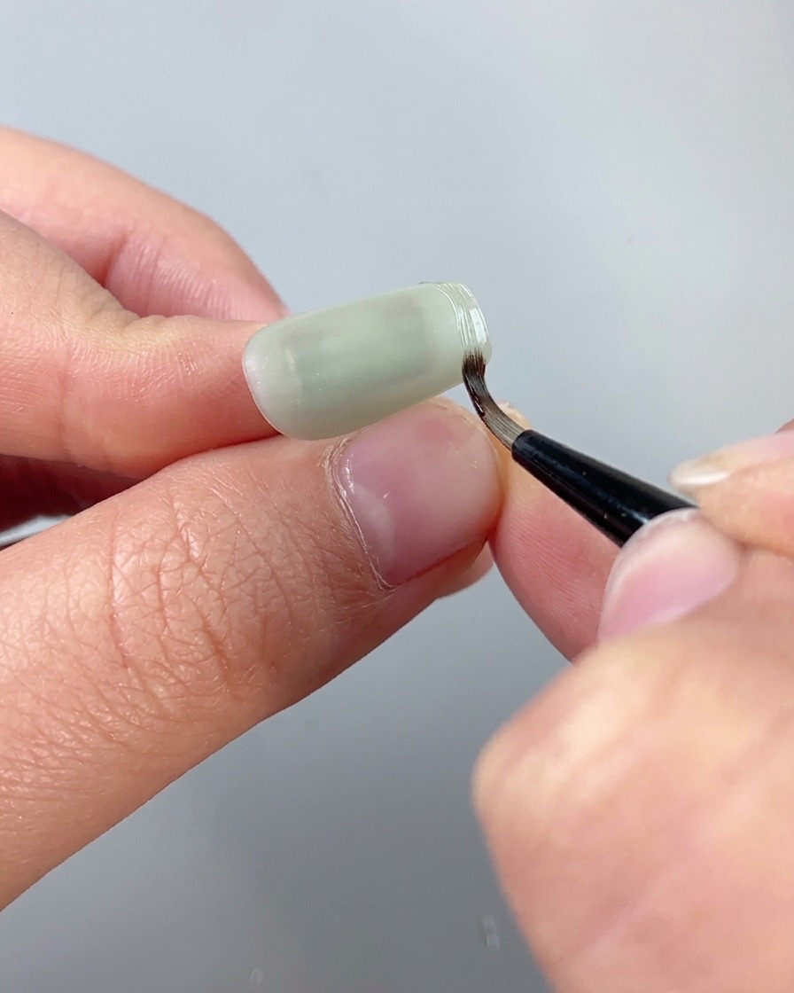 A fine detailing brush using clear texture sculpting gel to trace a raised border along the edge of a cat eye nail.