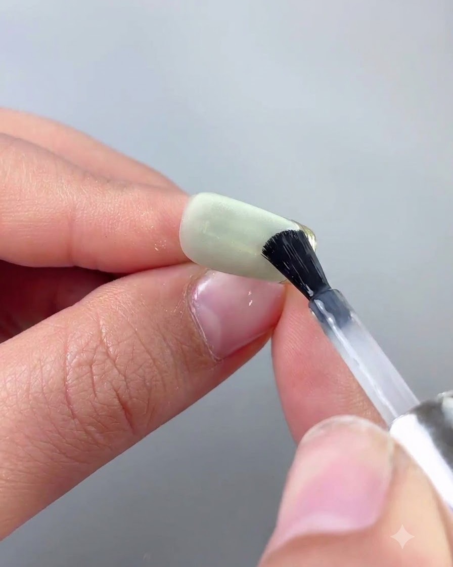 A fine nail art brush applying top coat precisely along a gold chrome edge of a green cat eye nail.
