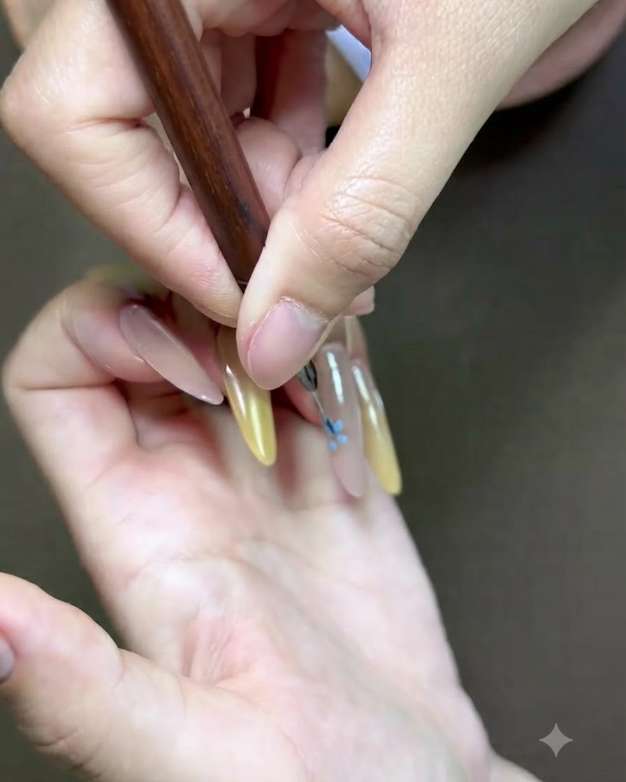 A detailed close-up of a fine nail art brush adding a pink floral accent onto the nail.