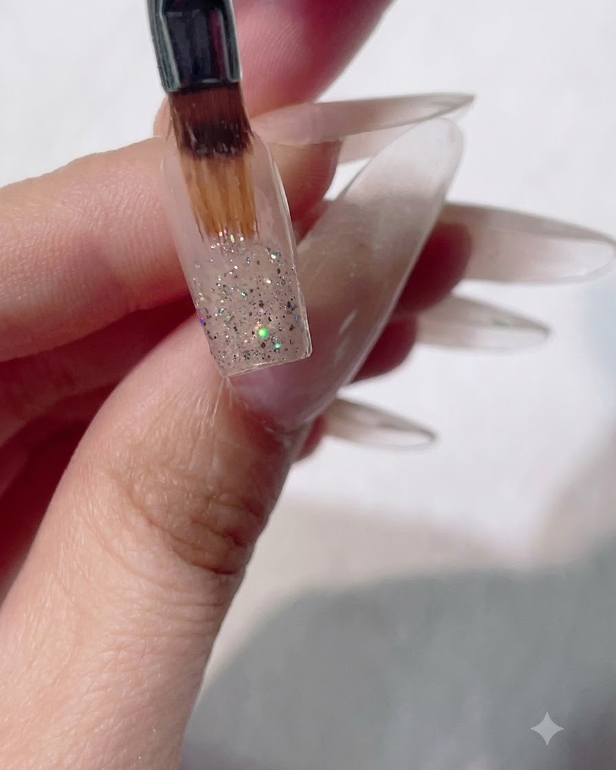 A flat gel nail brush sweeping wet glitter downward on a nail to create an ombre gradient effect.