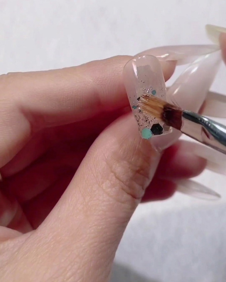 A small nail brush placing large blue and green hexagon sequins onto the wet glitter gradient on a fingernail.