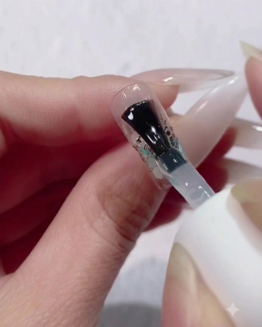 A bottle brush applying a thick, generous layer of clear builder gel over a glitter and sequin gradient nail design.