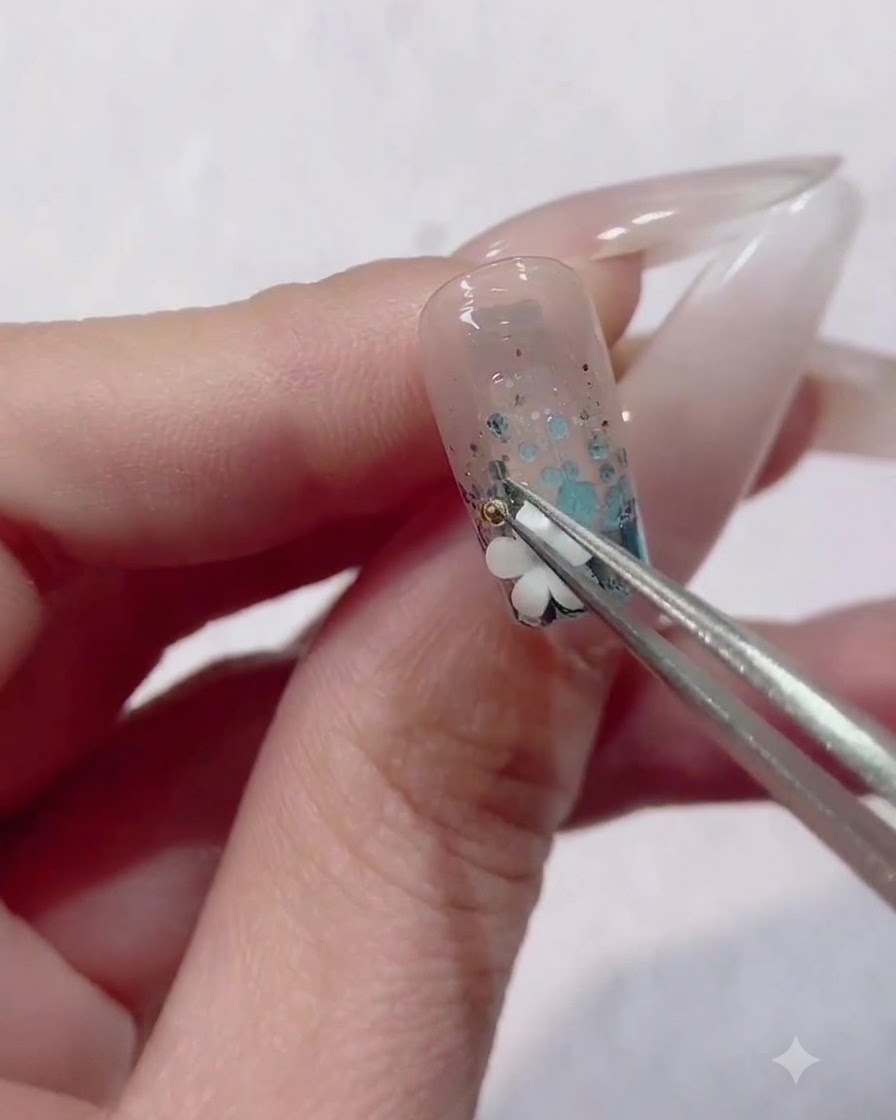 Precision tweezers placing a tiny metallic bead next to a white flower charm on a decorated nail.
